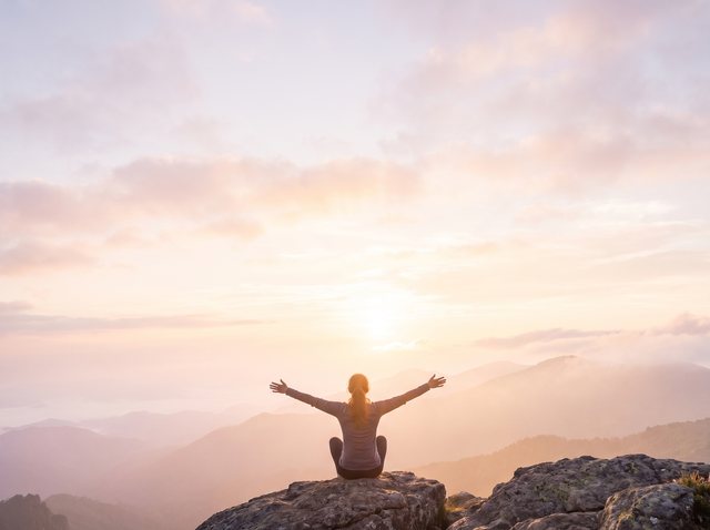 Eine Frau sitzt auf einem Felsen und öffnet die Arme zum Sonnenaufgang über den Bergen.