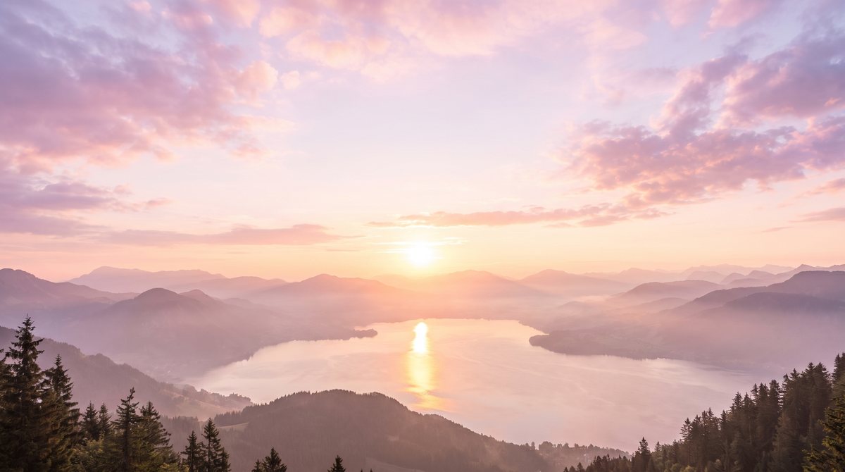 Sonnenaufgang über Bergketten und einem stillen See, der Himmel in Rosa und Gold.